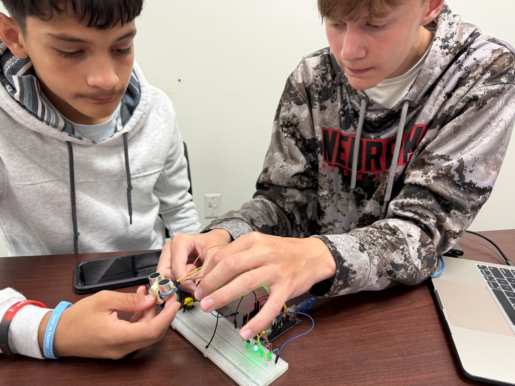 Two young boys are working on an electronics project with a breadboard, ultrasonic sensor, and LEDs. They are focused on connecting wires and building a circuit.