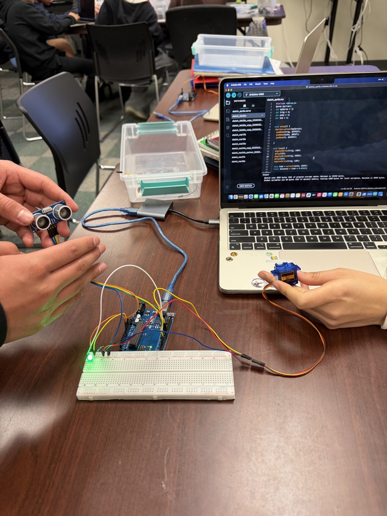 Two people are working on a robotics project, with hands holding an ultrasonic sensor and a servo motor connected to an Arduino board and a laptop displaying code. The setup is on a wooden table in a classroom environment.