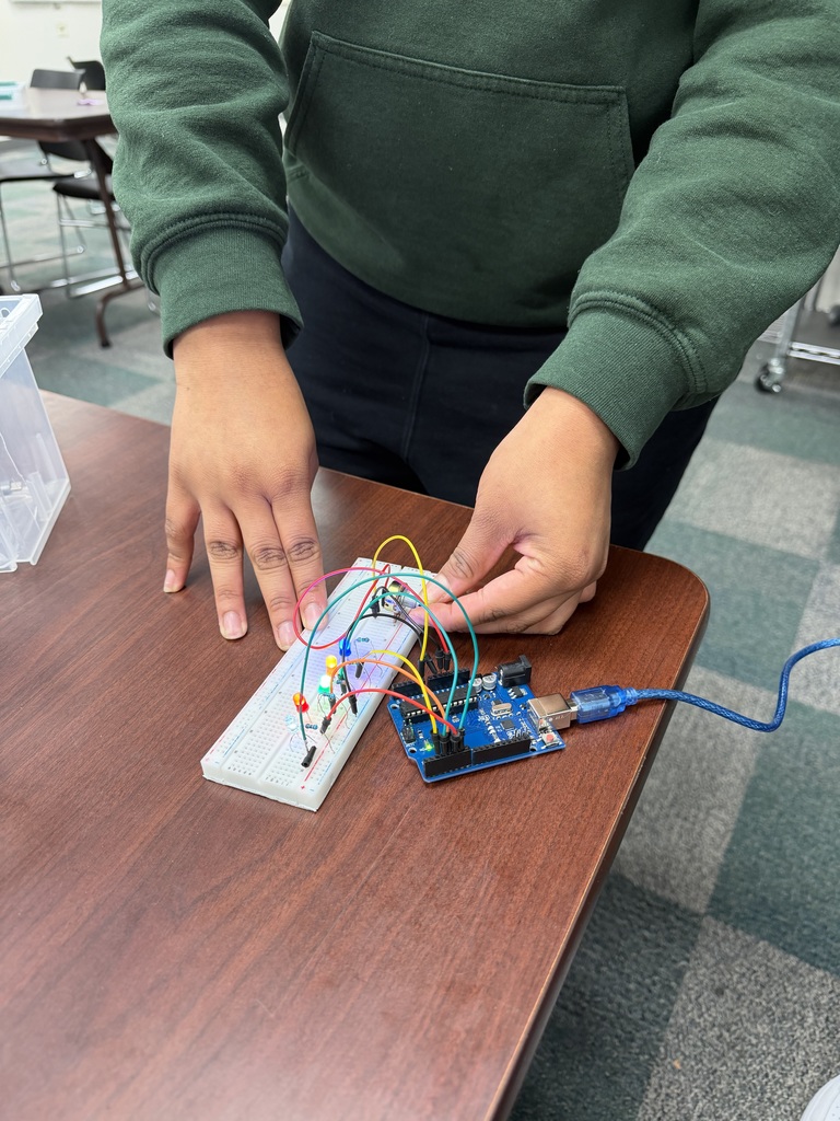 A person's hands are shown connecting wires to a breadboard with LEDs and an Arduino board. The setup is on a wooden desk.