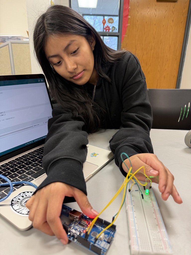 A young woman is focused on a electronics project, connecting wires between an Arduino board and a breadboard with lit LEDs, while referencing code on her laptop.