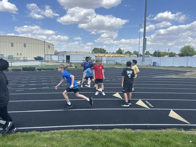 Several young boys are on a track, with one in the foreground running with a baton. They appear to be participating in a relay race or track practice on a sunny day.