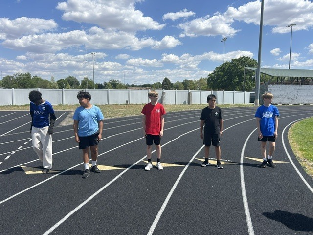 Five young boys are standing on a running track under a partly cloudy sky. They appear to be on the starting line of a race.