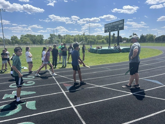 A group of children stands on a track field marked with numbers, likely preparing for or participating in a track and field event. An adult coach or official observes them, while others watch from the sidelines under a partly cloudy sky.
