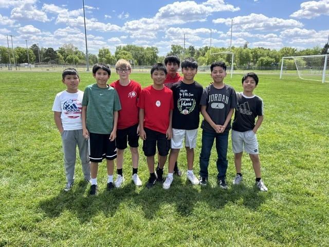 A group of eight young boys are standing on a grassy field in front of soccer goals. They are all looking towards the camera and appear to be posing for a photo.