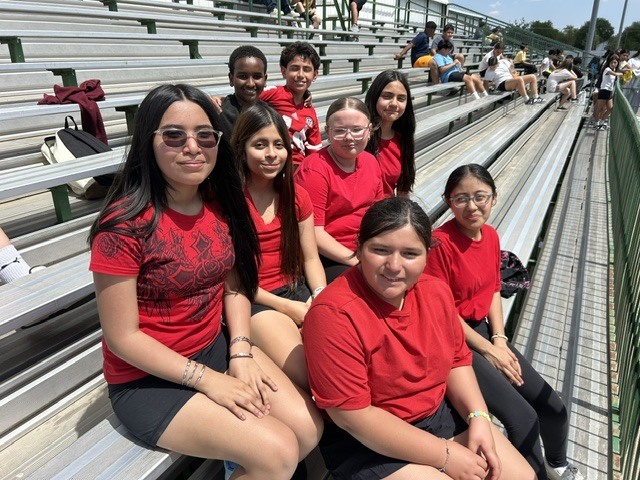 A group of young people wearing red shirts are sitting on bleachers. They appear to be at an outdoor event like a sporting competition.