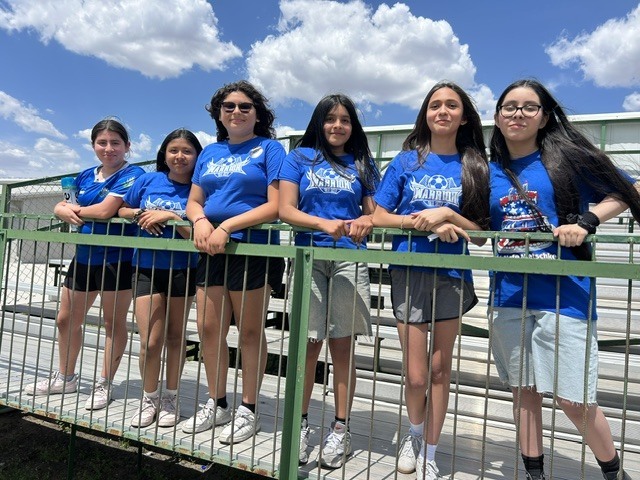 Six young girls in blue t-shirts stand behind a green railing at a sporting event. They are all looking towards the camera on a sunny day.