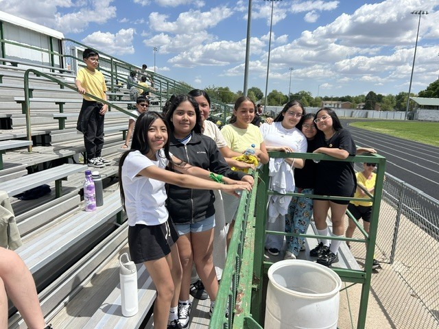 A group of young people are posing for a photo on stadium bleachers on a sunny day. They appear to be at a sporting event, possibly a track meet.