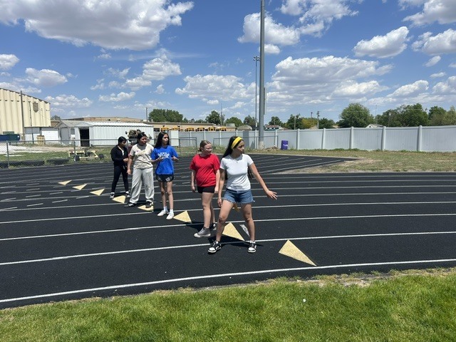 A group of young women stand on a running track, some looking towards the viewer. The scene is outdoors on a sunny day with a clear blue sky and scattered clouds.