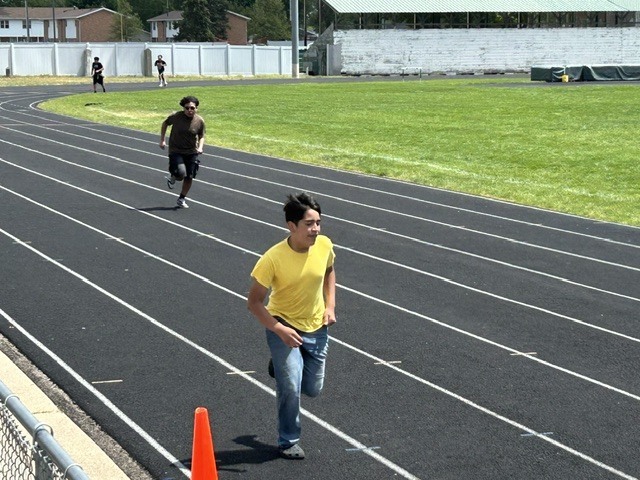 Several young people are running on a black track with white lane markers. In the foreground, a boy in a yellow shirt runs towards the viewer.
