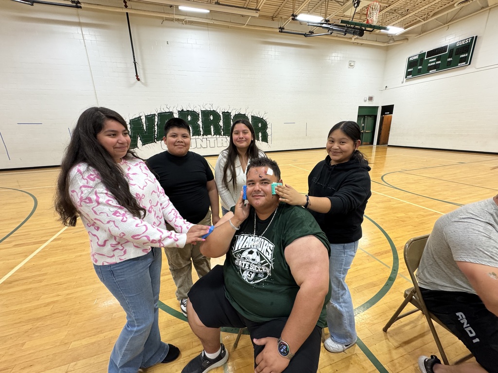 In a school gymnasium, a group of students surround a seated teacher, playfully applying temporary tattoos to his cheeks while he smiles. Behind them, a large “WARRIOR” mural and basketball hoop indicate the setting is likely a school sports facility.