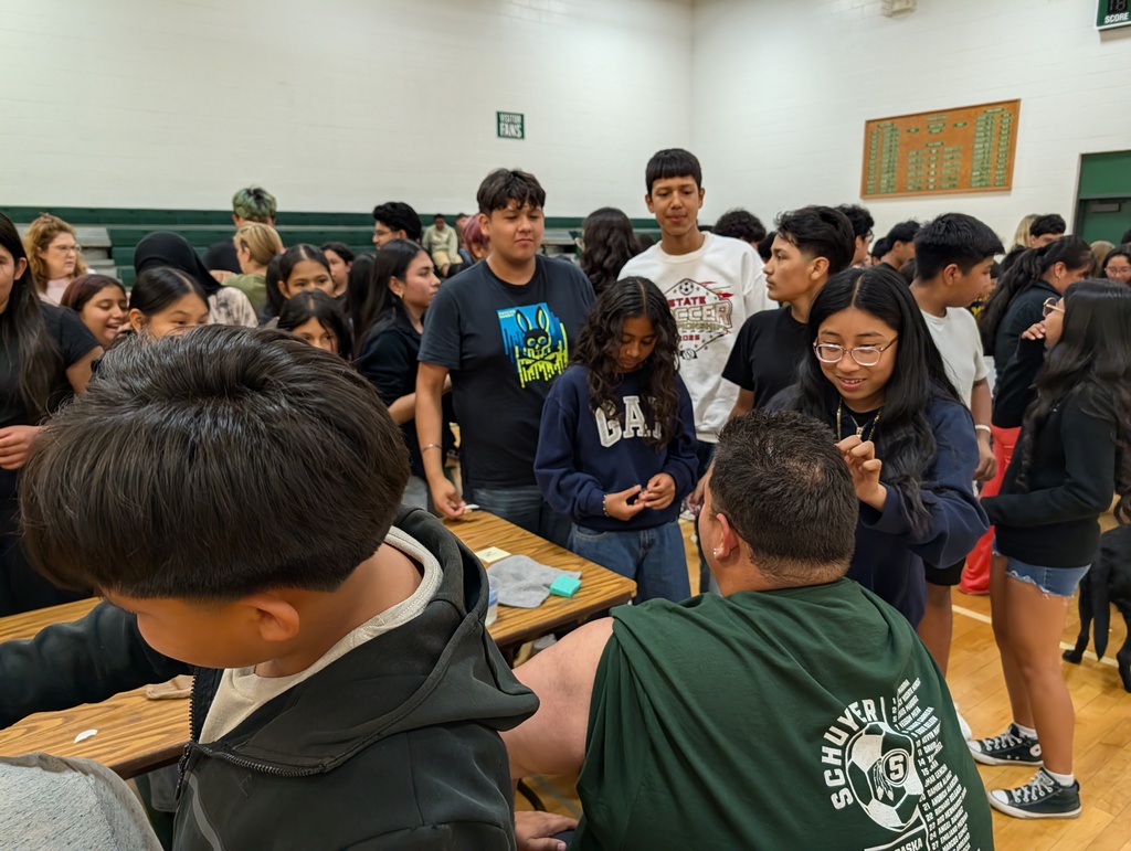 A group of young people are gathered in a gymnasium, waiting to apply a temporary tattoo to a teacher in a green shirt.
