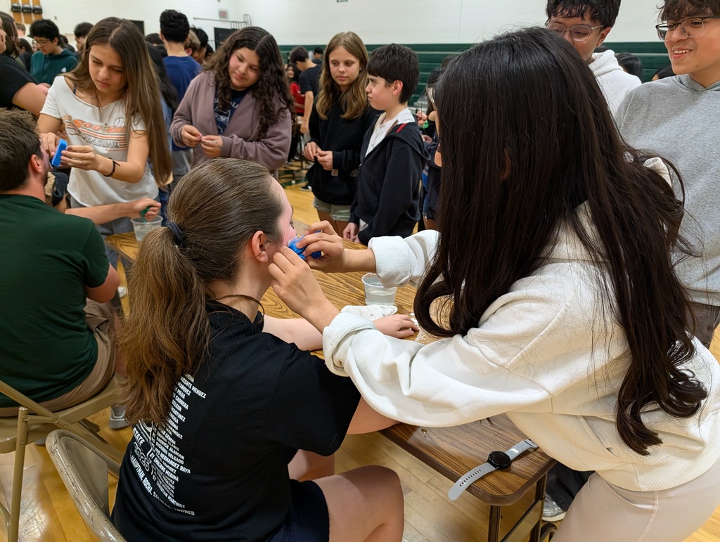 A young person in a white hoodie applies a temporary tattoo to a teacher's cheek. Other young people stand around them in a gymnasium waiting their turn..