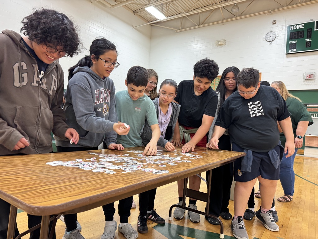 A group of students gathers around a table in a gymnasium, actively searching through tempory tattoos to put on their teacher.