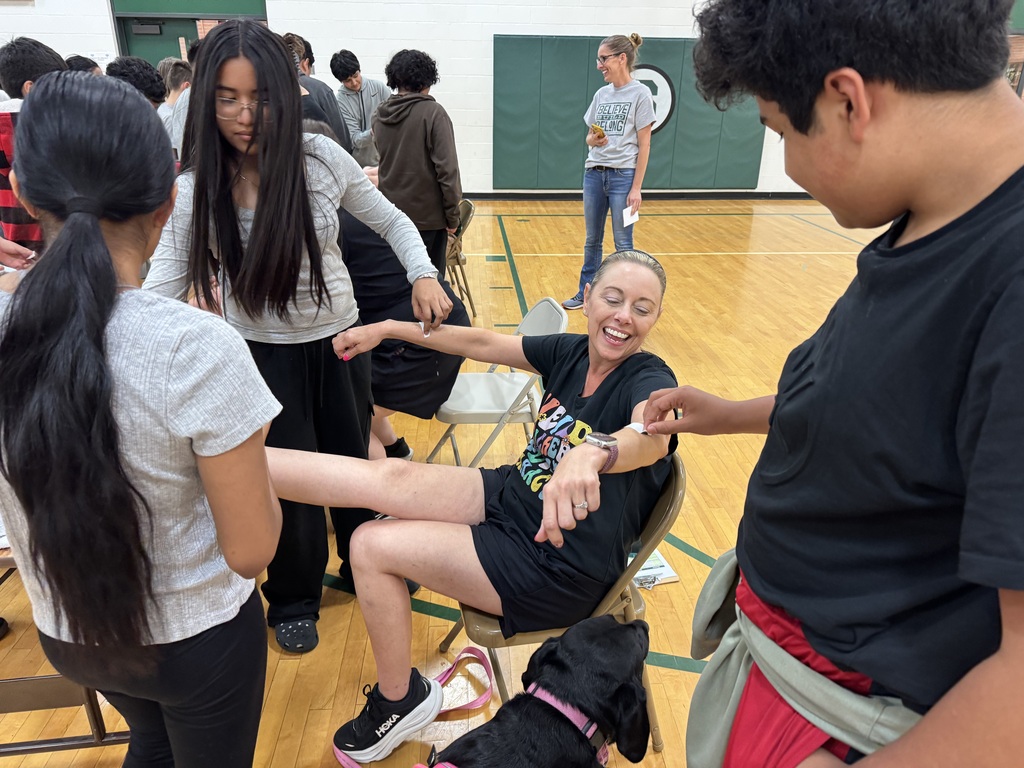 A smiling woman sits in a chair in a gym while several young people, a dog, and a teacher stand around her. The young people are placing tattoos on the woman's arms and leg