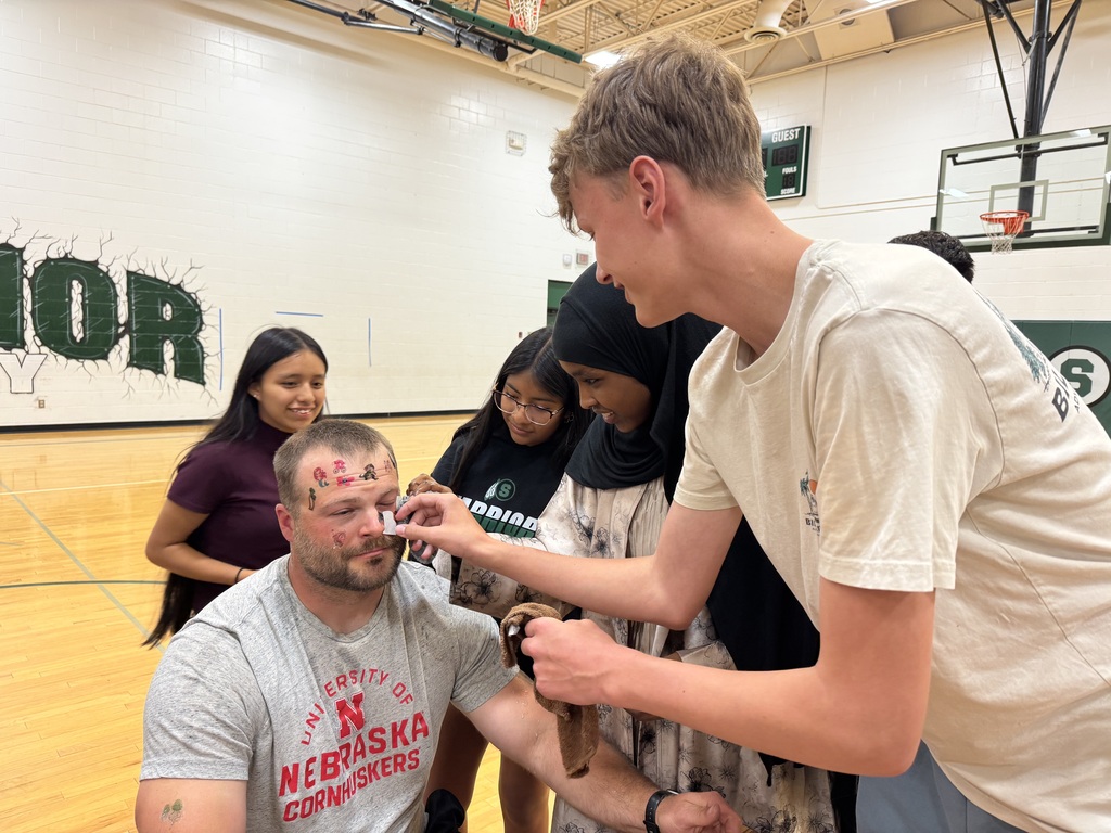 A teacher wearing a Nebraska Cornhuskers t-shirt is getting temporary tattoos applied to his face by a group of young people in a gymnasium. The people appear to be enjoying themselves and the man has a relaxed expression.