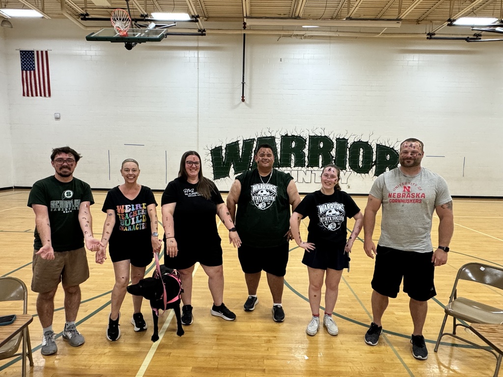 Six people with tatoos and a black dog with a pink leash are posing together on a gymnasium floor, all covered in paint splatters. Behind them, a large "WARRIORS" mural is visible on the wall, along with an American flag and a basketball hoop.