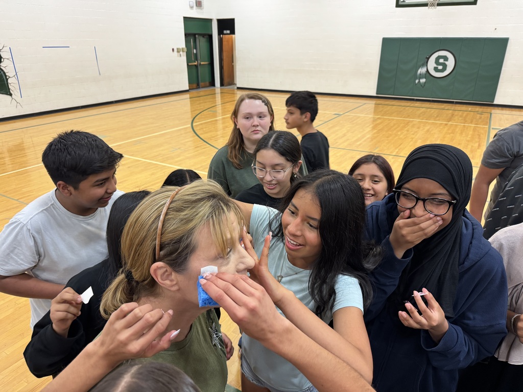 A group of students are gathered around a teacher in a gymnasium, appearing to be applying a temporary tattoo to her face. The students are smiling and engaged in the activity.