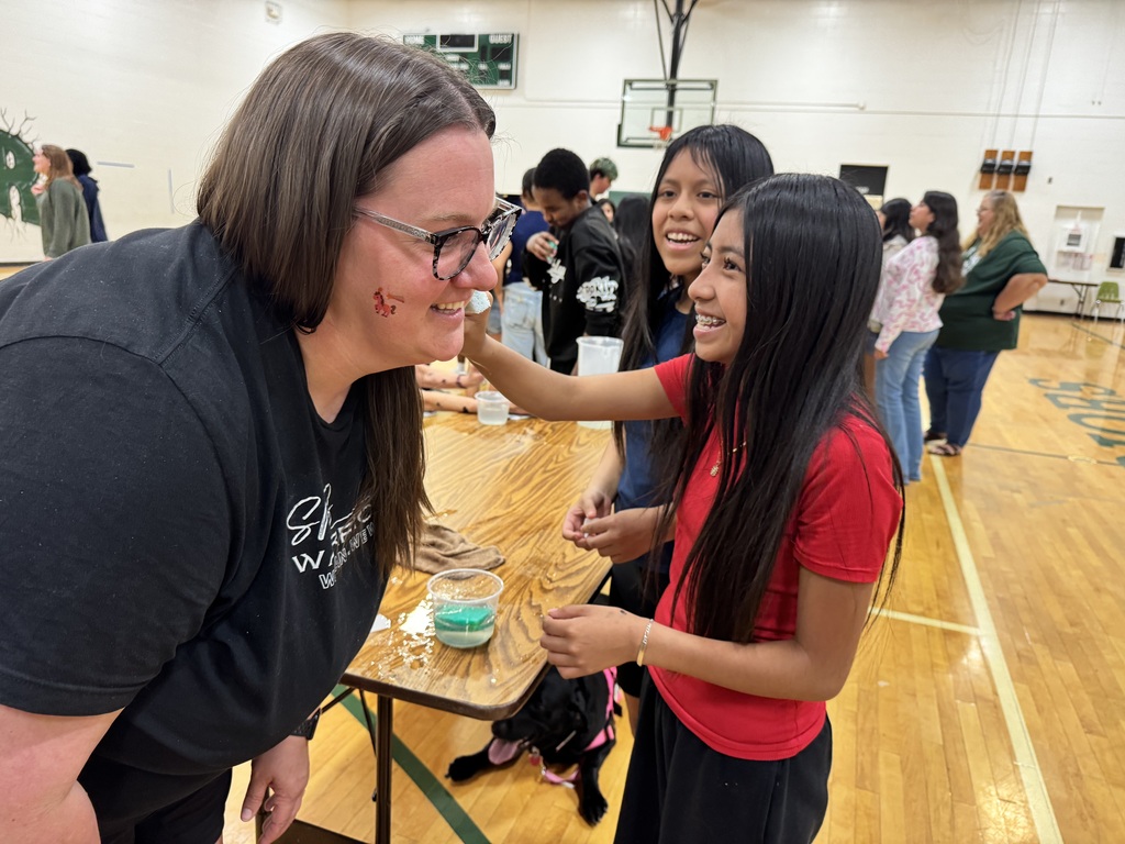 A girl in a red shirt is applying a temporary tattoo to the cheek of a teacher wearing glasses. They are in a gymnasium with other people and a black dog nearby.