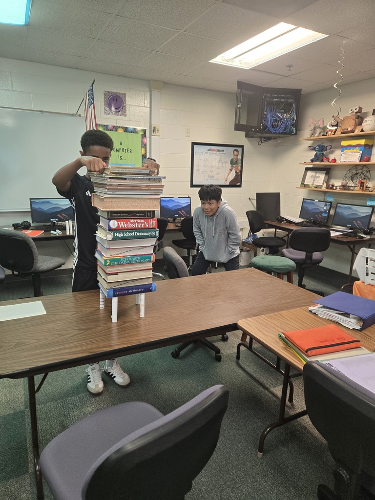 The photo shows a classroom where two students are participating in a fun activity. One student is balancing a tall stack of books on a small stand, while another watches nearby. Computers, classroom supplies, and desks fill the background.