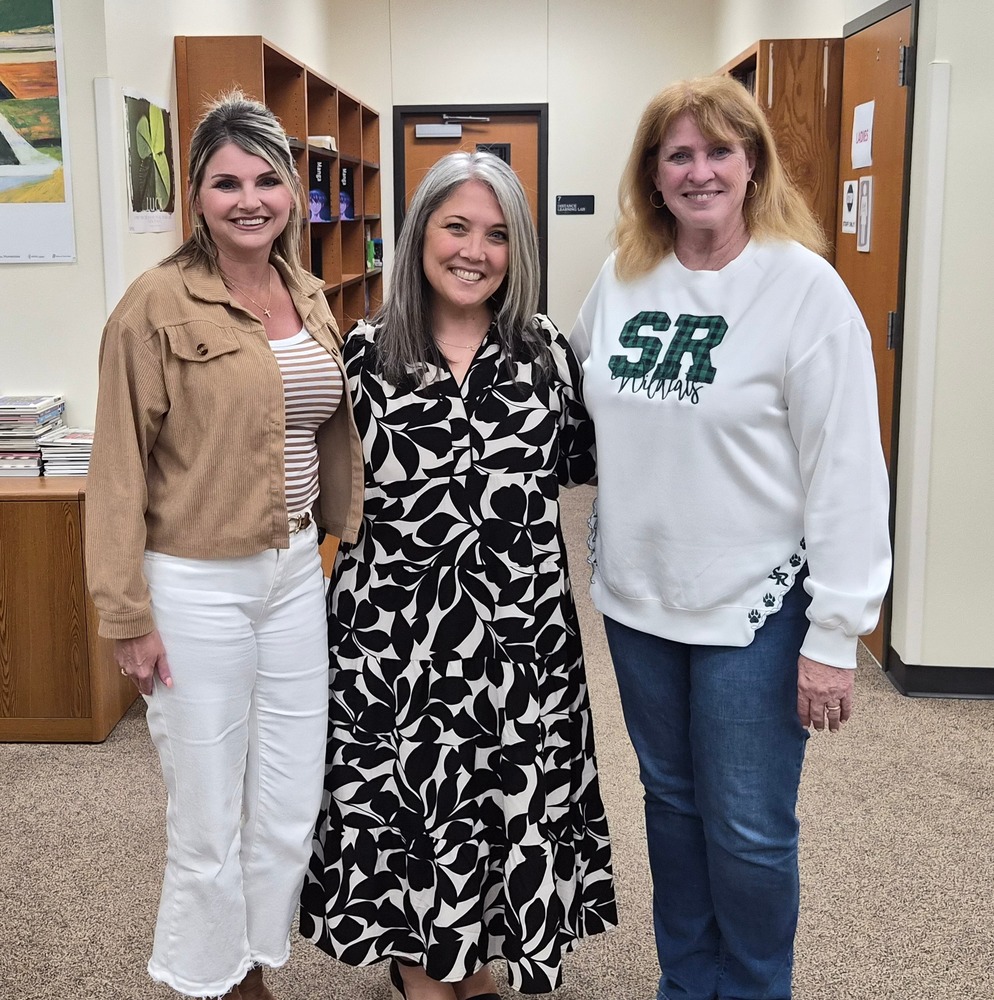Three women stand side by side in a school hallway, smiling at the camera. The woman on the left wears a tan jacket over a striped top with white pants; the woman in the middle wears a black-and-white patterned dress; and the woman on the right wears a white sweatshirt with green “SR Wildcats” lettering and blue jeans. Behind them are wooden shelves, a door, and wall artwork, suggesting an office or library area.