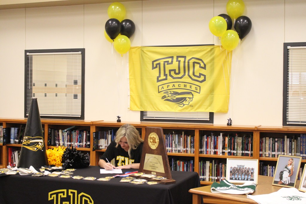 teenage blonde student sitting at a table with a black table cloth sign her college committment letter