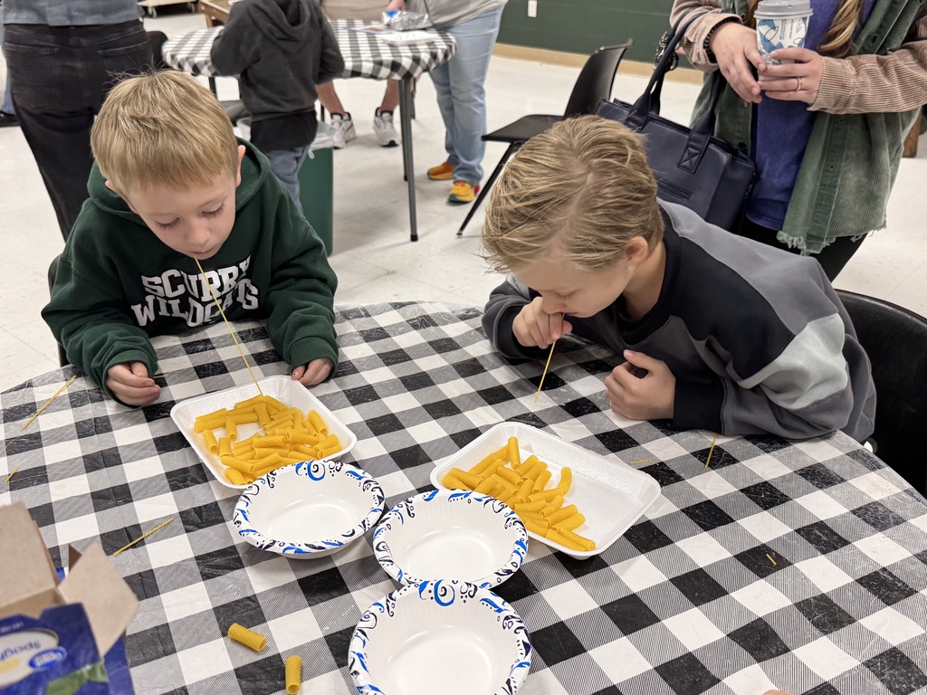 two male students playing a game.  have to use a stick to pick up the most pasta without hands