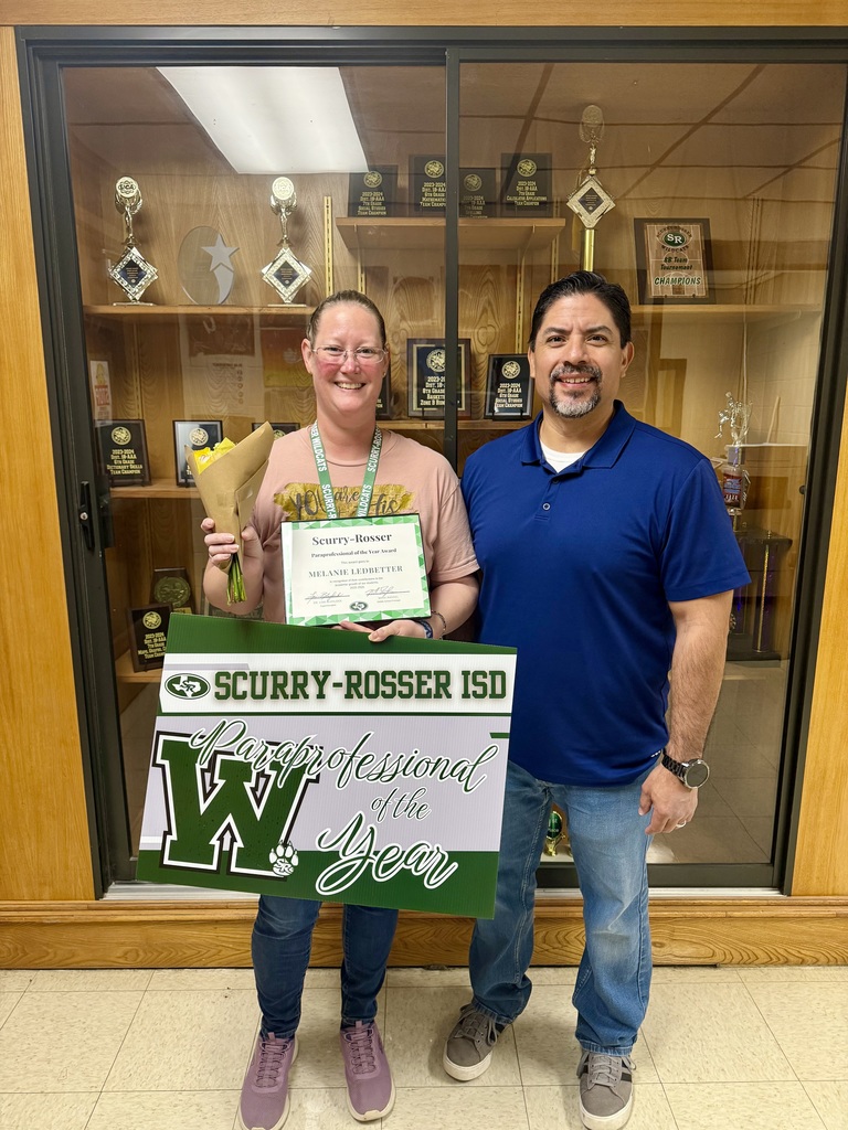 picture of a man in a blue collared shirt standing next to a women holding a sign in front of a trophy case