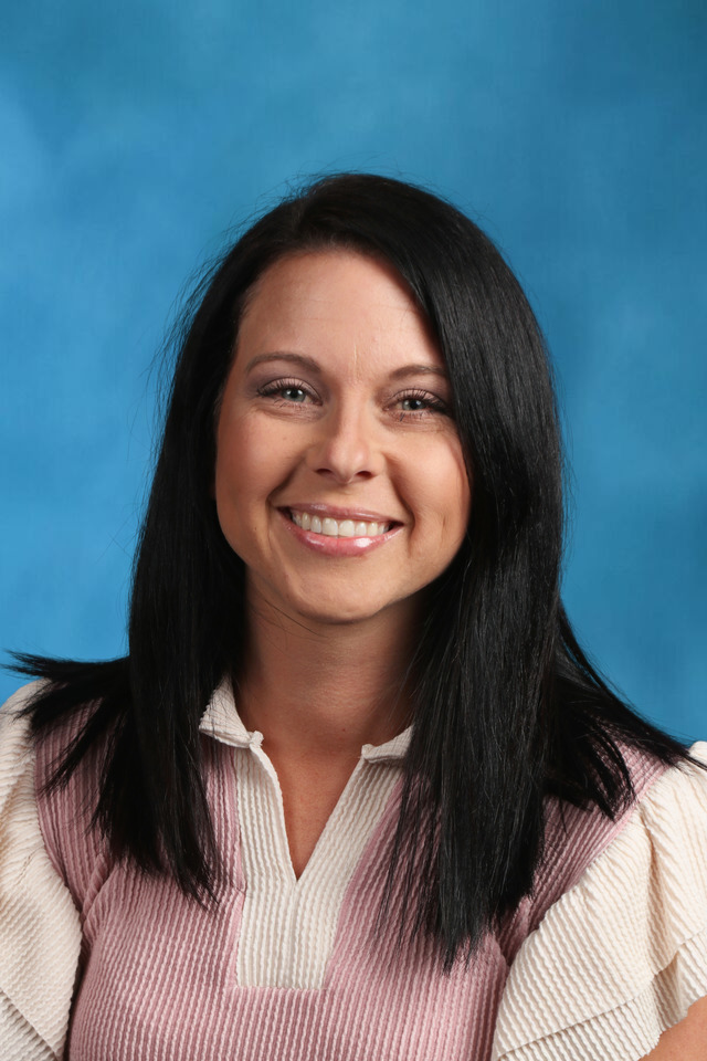 Head-and-shoulders portrait of a woman with straight, dark shoulder-length hair, smiling at the camera against a solid blue studio background. She wears a light pink textured top layered over a cream blouse with a ruffled collar, giving a polished, professional appearance.