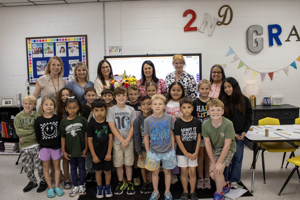 Group photo in an elementary classroom showing teacher and five members of leadership standing behind a large group of young students, all smiling at the camera. 