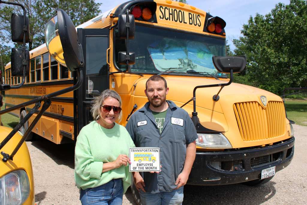 female bus driver standing with male supervisor in front of a yellow bus being names bus driver of the month