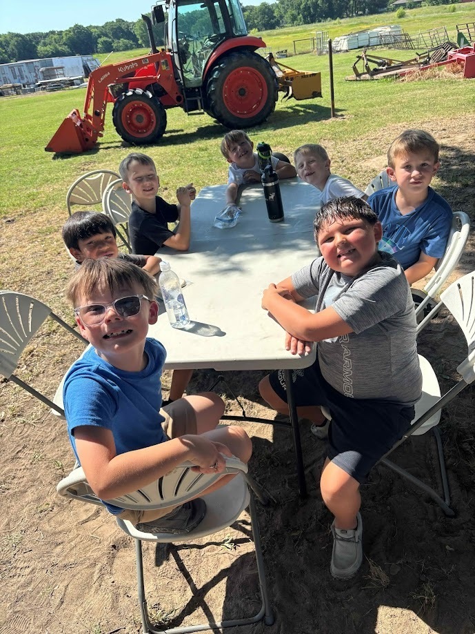 a group of male students sitting at a plastic table outside with a tractor in the background
