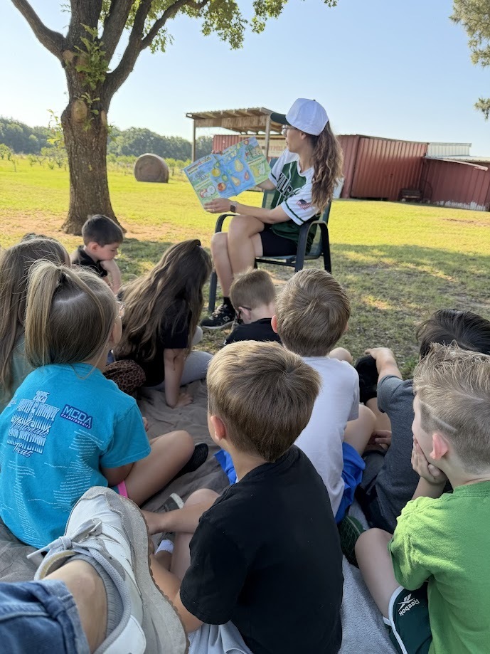 group of students listening to an adult worker read a book while sitting in the grass under a tree.