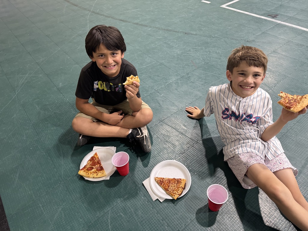 two male students sitting on the gym floor eating pizza