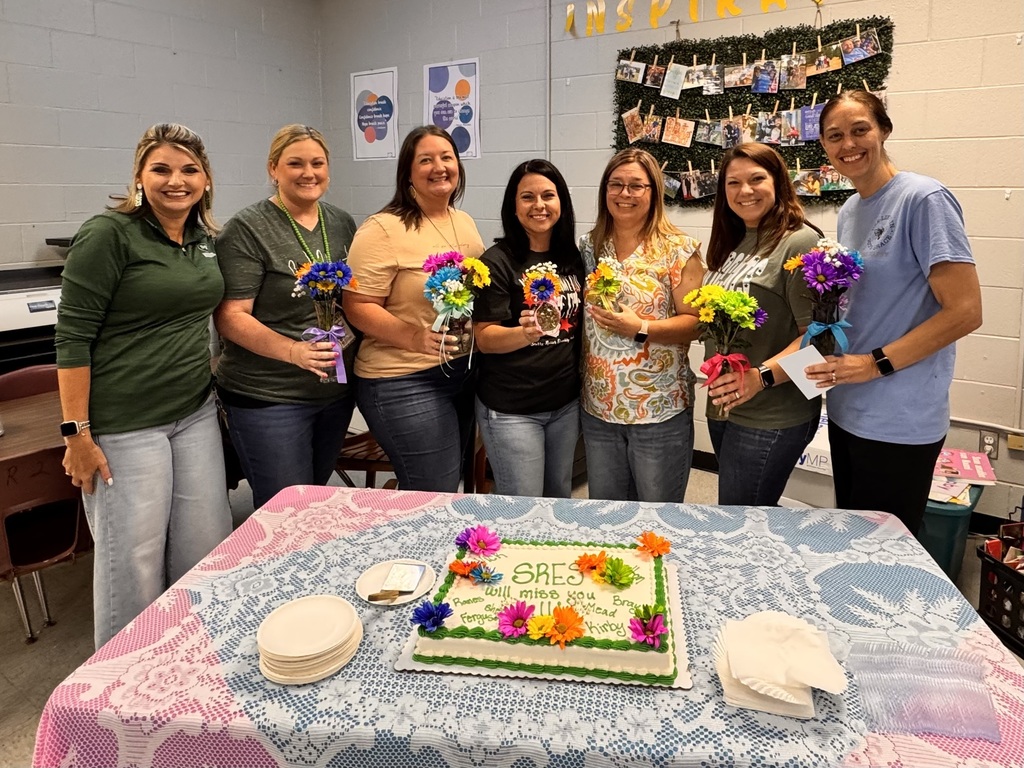 7 ladies holding flowers standing in front of a decorated cake