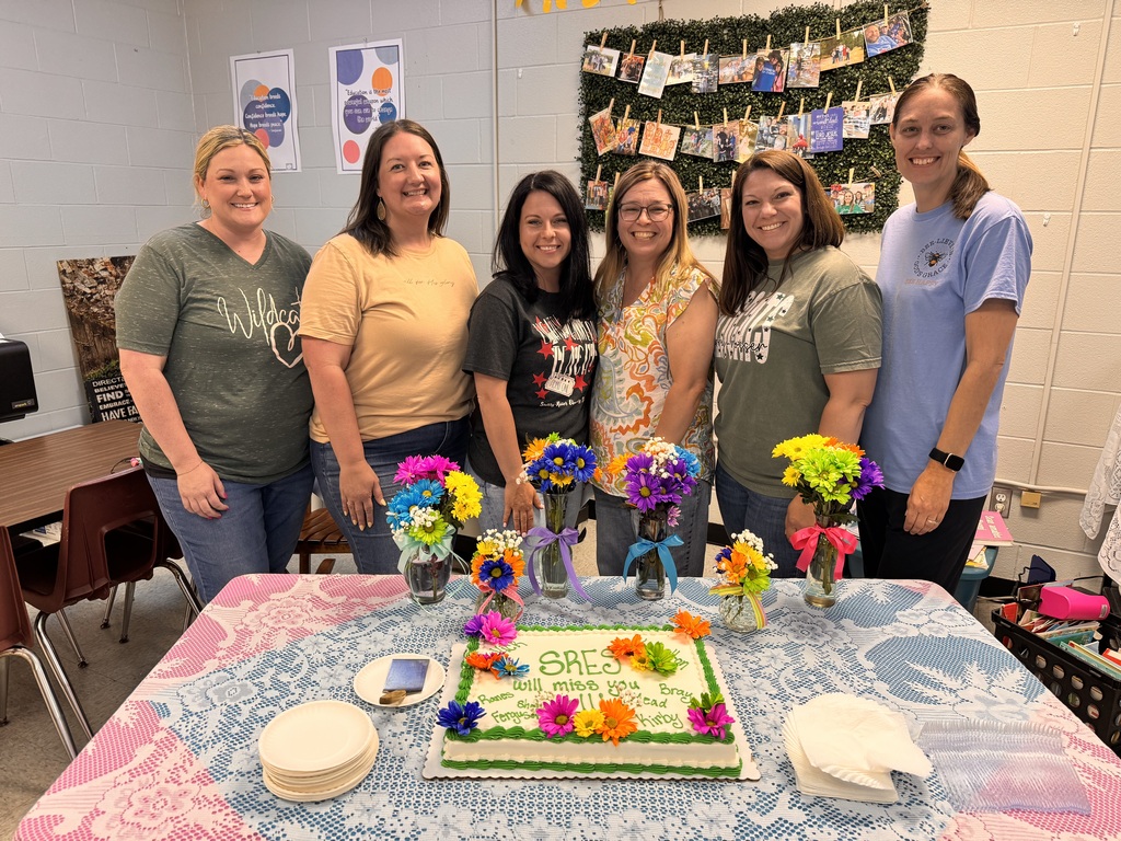 6 ladies standing in front of a table with a decorated cake and vases of flowers
