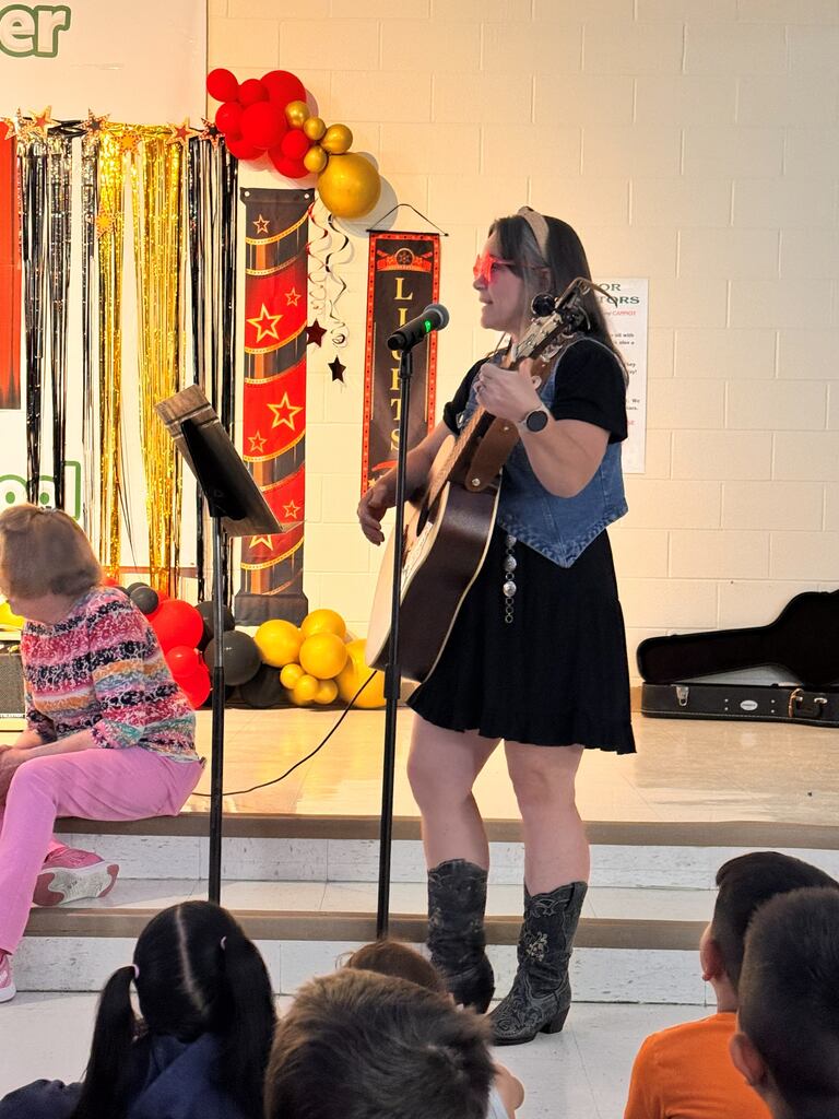 female teacher wearing a black dress and denim best playing the guitar and wearing start glasses