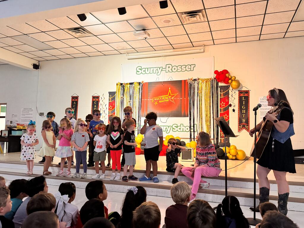 group of prek students wearing glasses while singing on stage with their teacher playing the guitar and her assistant sitting on stage next  to another student