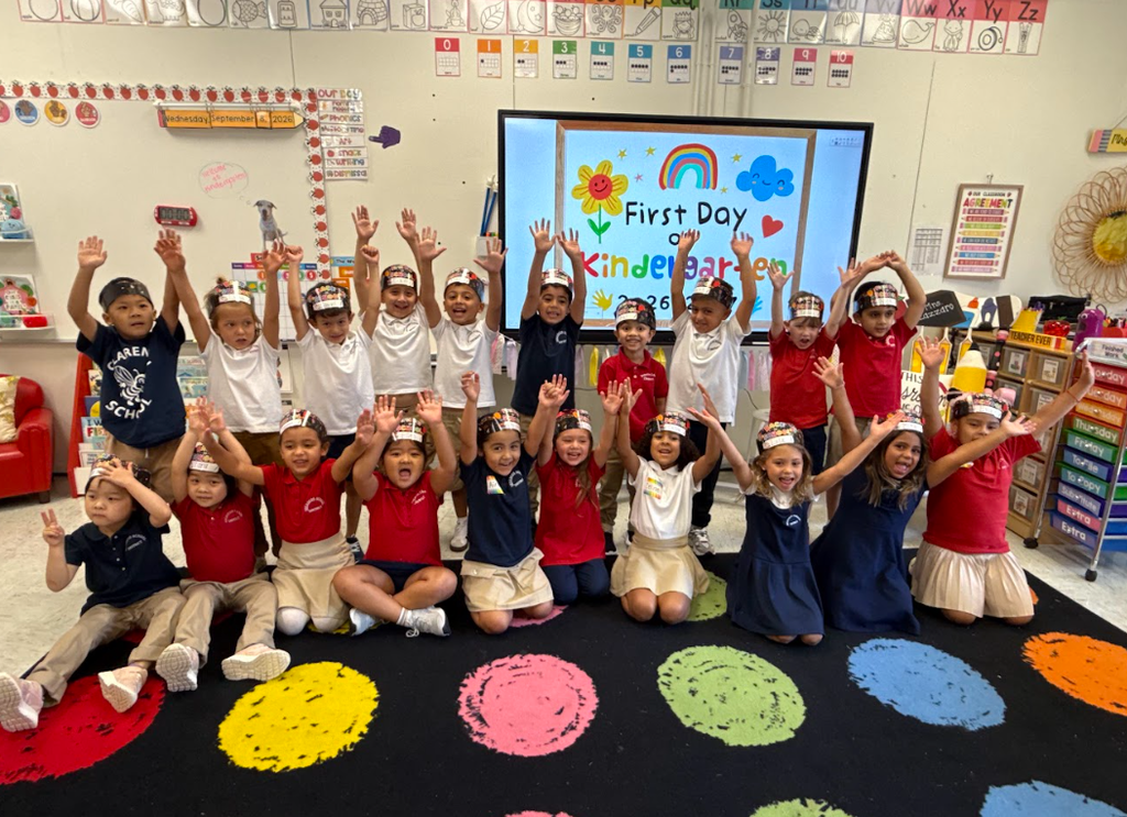 Classroom filled with students, all wearing hats, sitting or standing on a colorful floor mat. A screen in the background displays "First Day Kindergarten".