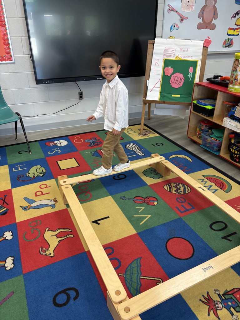 A young child wearing a white long sleeve shirt, pants, and white shoes walking on a wooden plank.