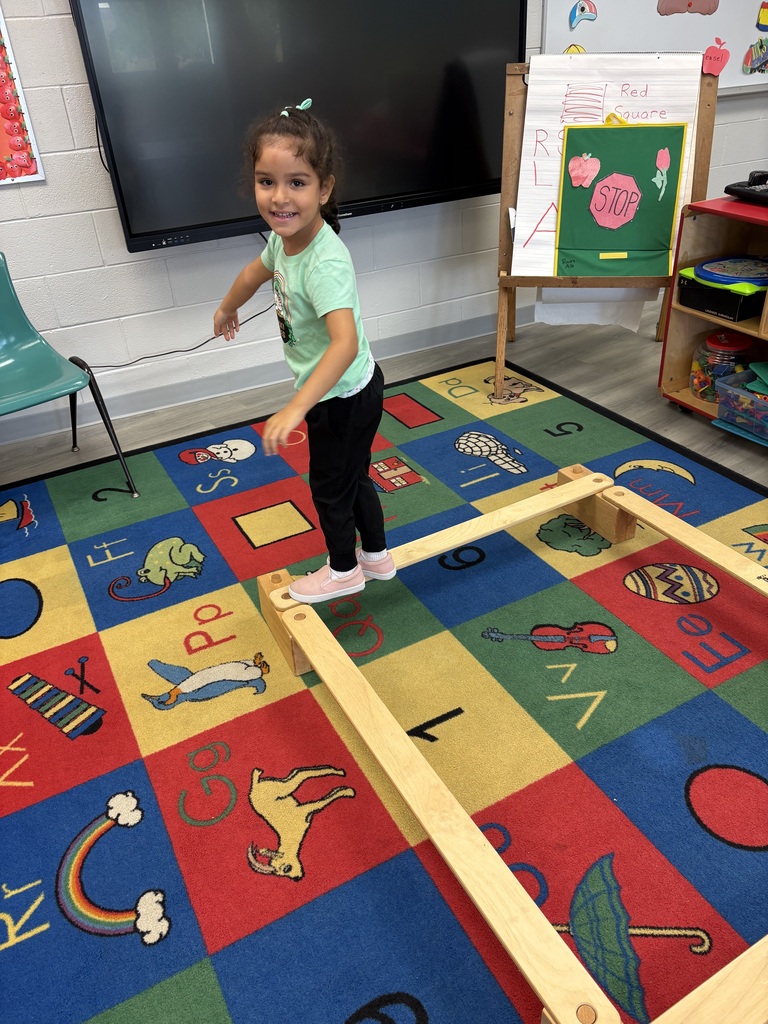 Child standing on wooden balance beam with alphabet and shapes rug on floor. Bright wall with framed art.