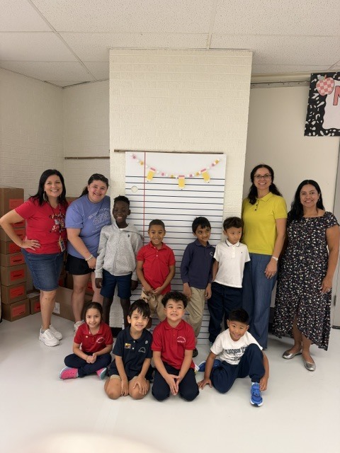 Nine children and three women pose for a photo in a classroom with a whiteboard, boxes, and a bulletin board.