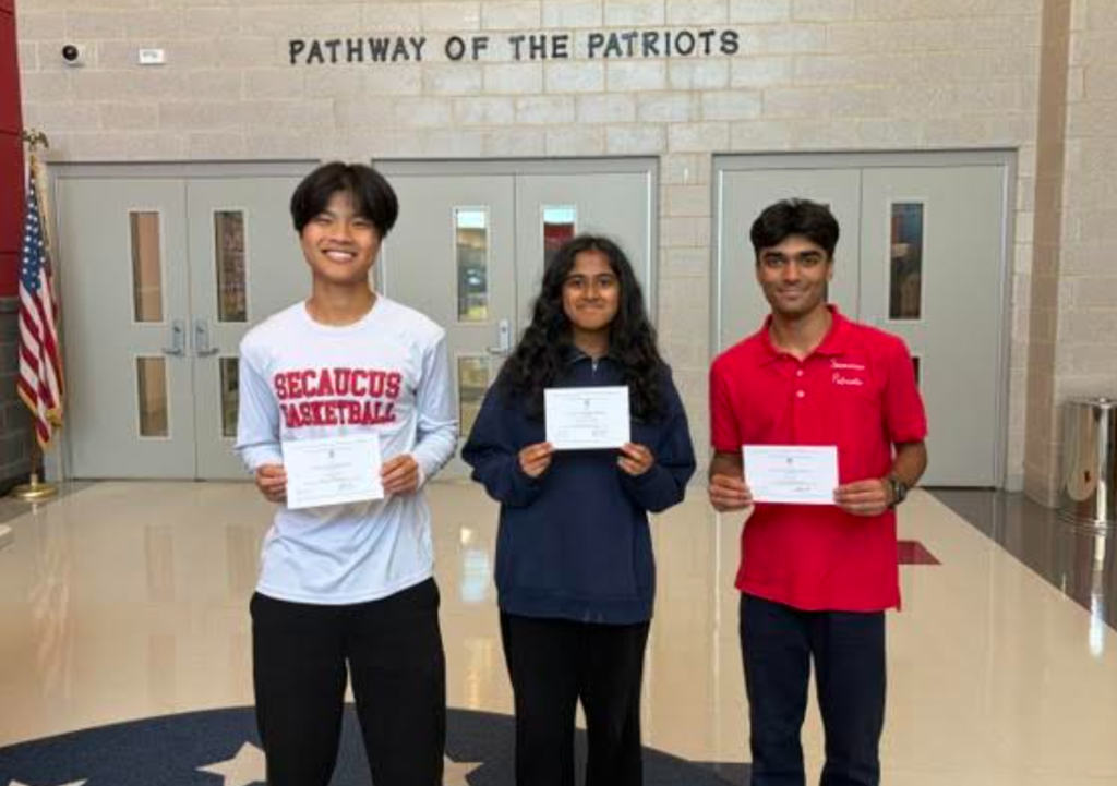 Three individuals pose in a hallway with a sign reading "Pathway of the Patriots". They hold certificates.