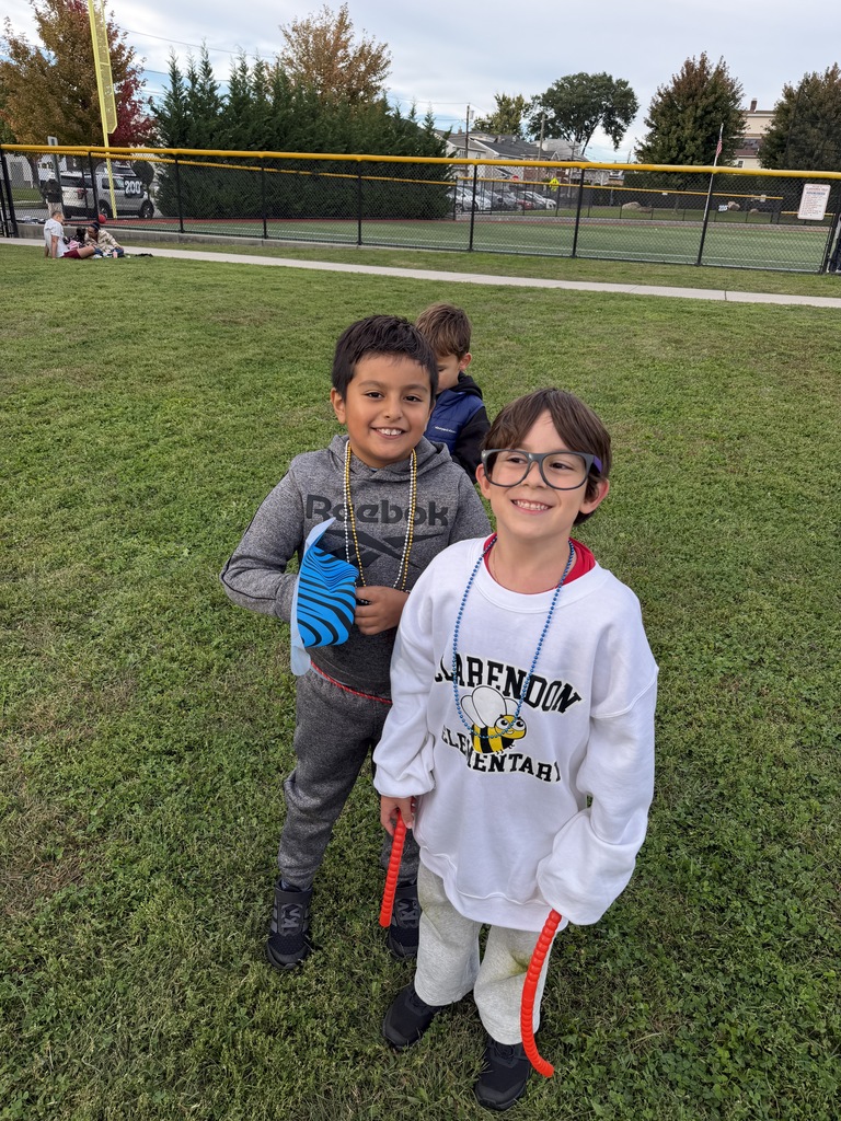 Two boys stand on a grassy field. One wears a white shirt with a logo. They stand near a fence with a tennis court in the background.