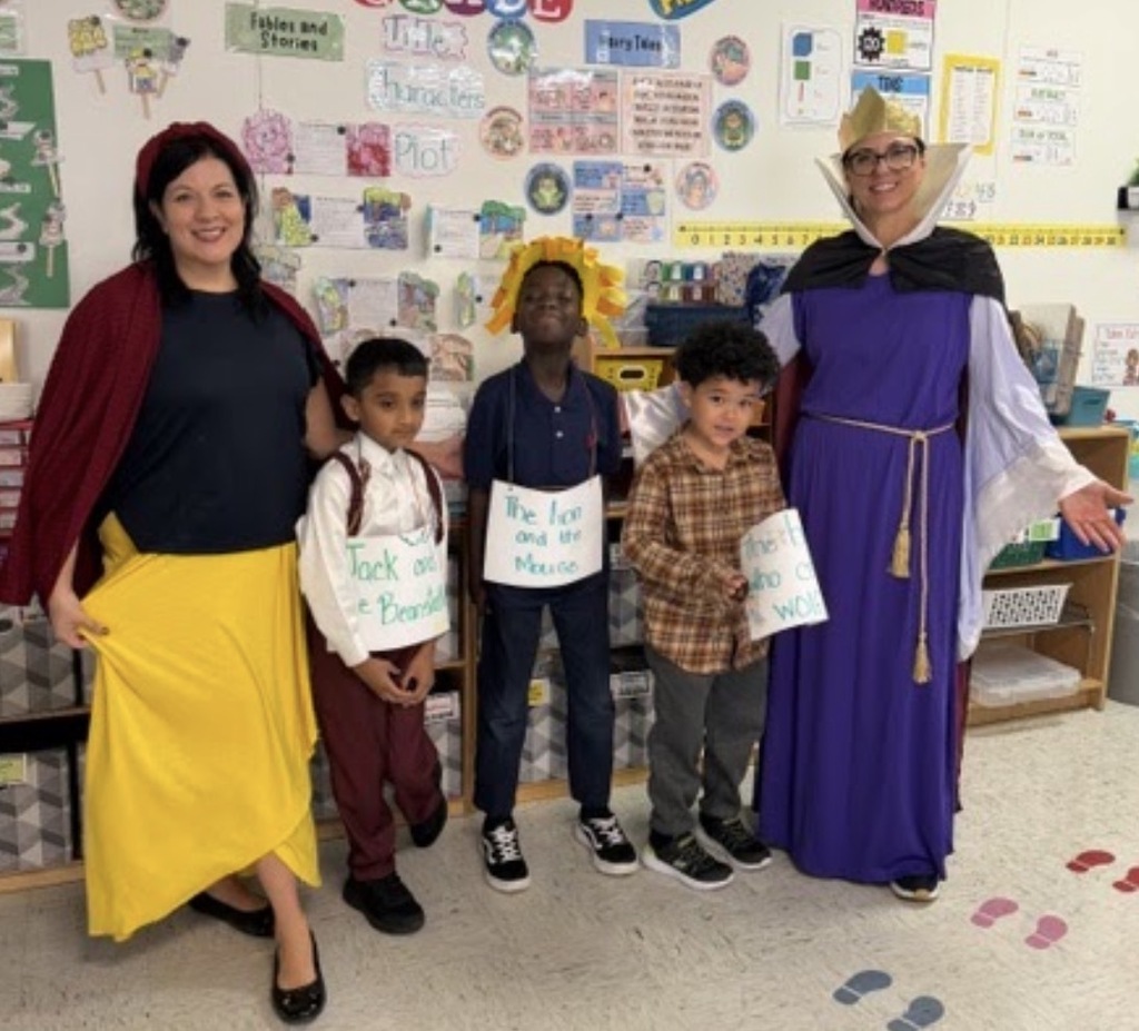A group of adults and children stand in a classroom, all dressed in costumes. One woman wears a cape.
