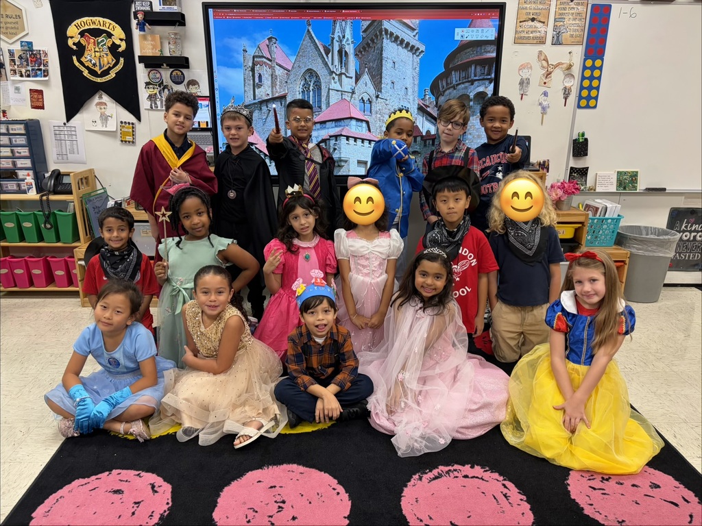 Kids in costumes pose for a group photo in a classroom with a large screen in the background.