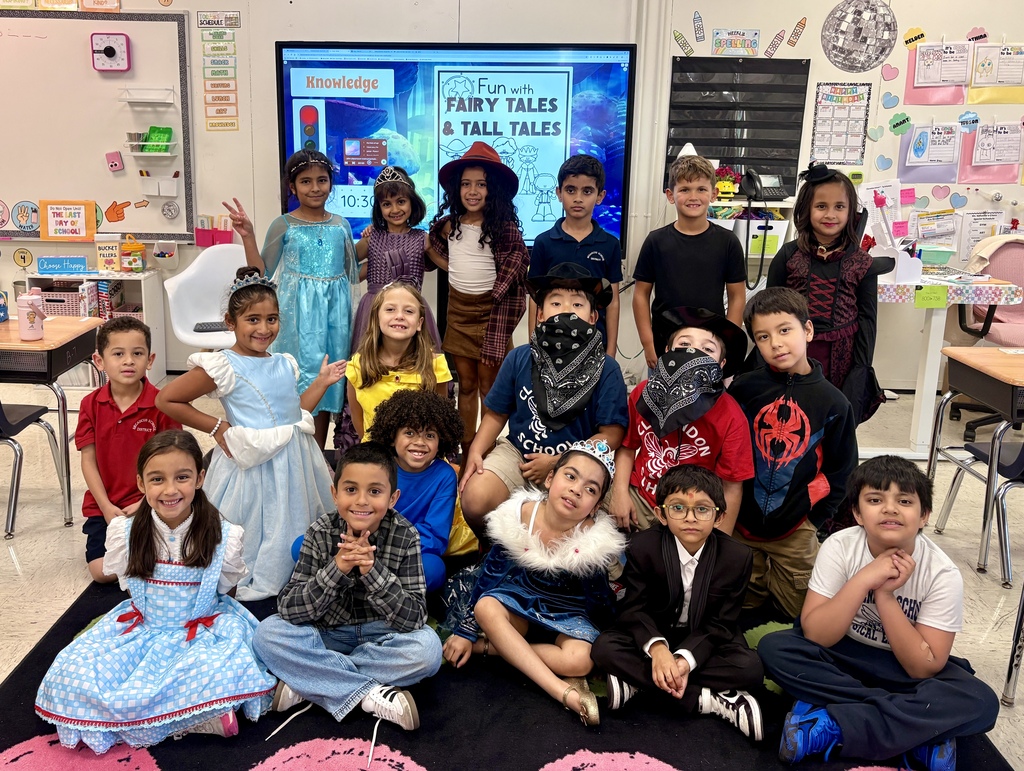 Students dressed in costumes sit on a rug in a classroom with a screen showing "Fairy Tales & Tall Tales."