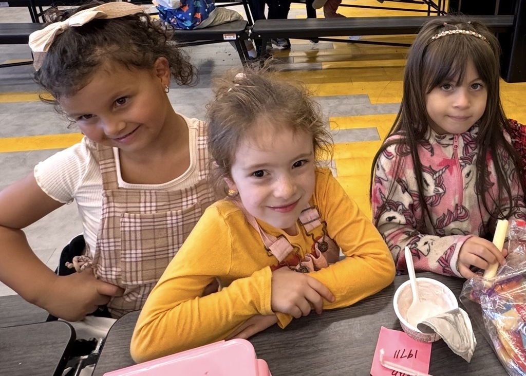 Three girls in a school cafeteria, seated at a table. One girl in a yellow shirt smiles.