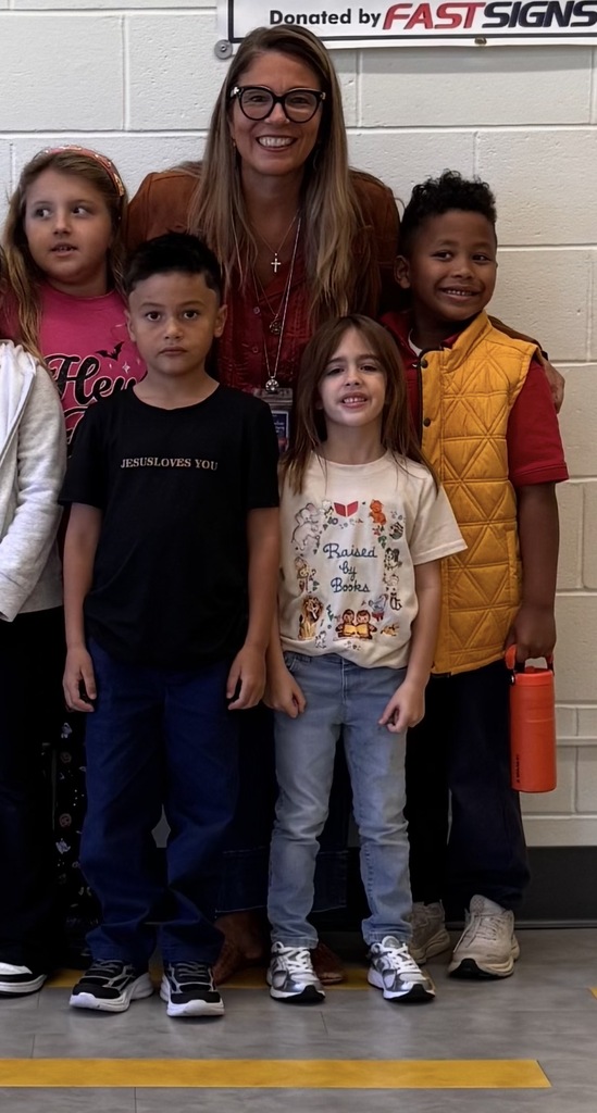A group of children stands with a woman, likely a teacher, in a school hallway. Behind them is a brick wall with a sign reading "Donated by FASTSIGNS."
