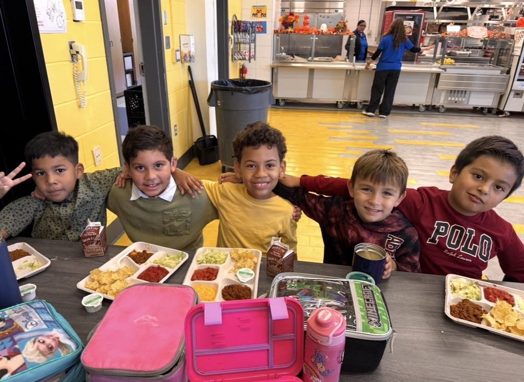Five children seated at a table, smiling for a photo. Trays of food, cups, and lunchboxes are on the table.