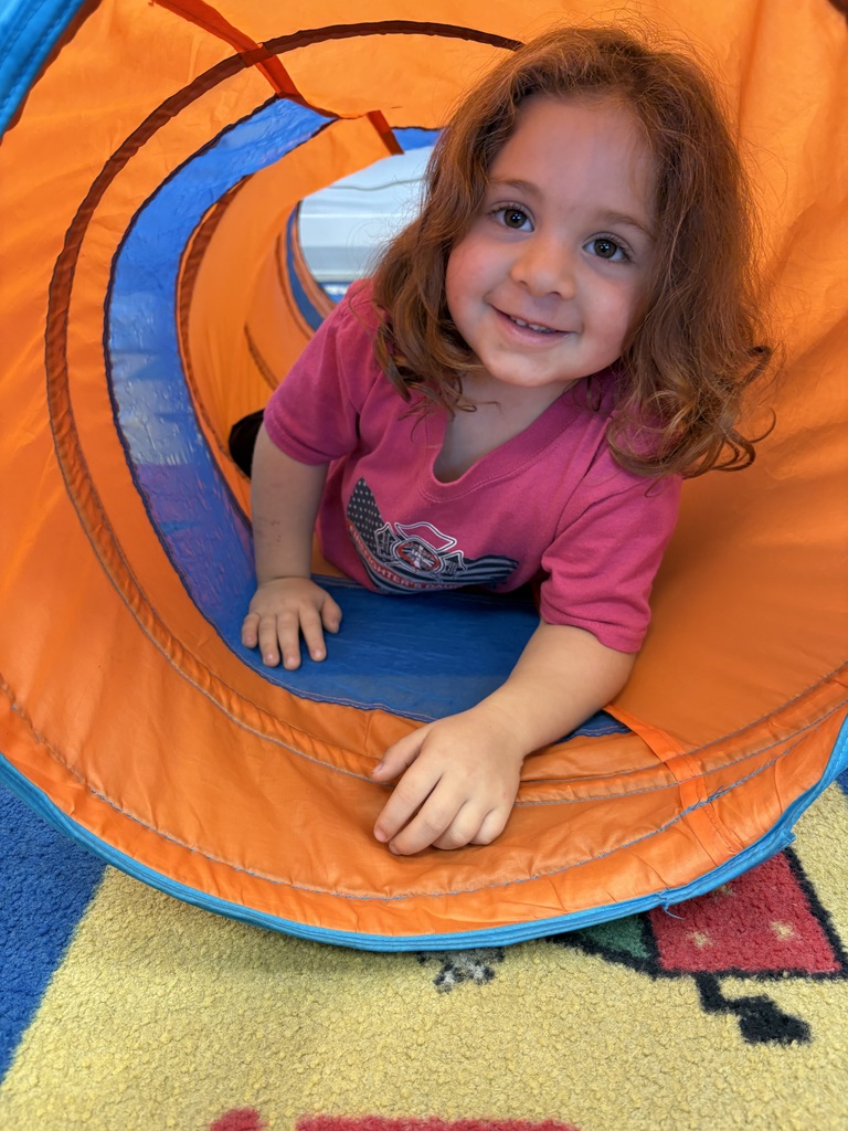 A young girl wearing a pink shirt crawls through a spiral orange and blue play tunnel on a carpeted floor.